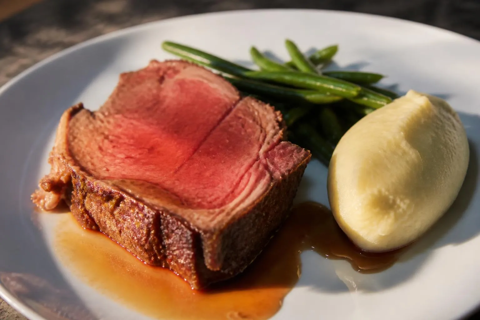 Une tranche de rôti de bœuf saignante à côté d’une purée et de haricots verts, sur une assiette blanche