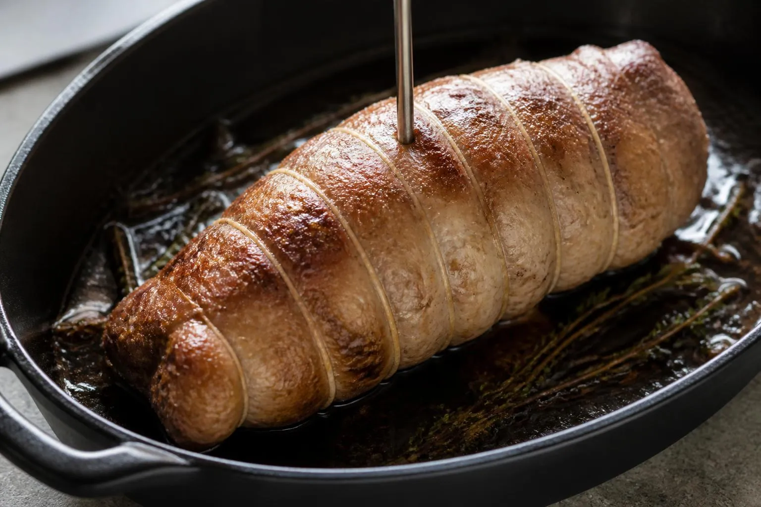 Un filet mignon de porc ficelé dans un plat à four en fonte, doré sur toutes ses faces, avec un thermomètre à sonde planté au centre